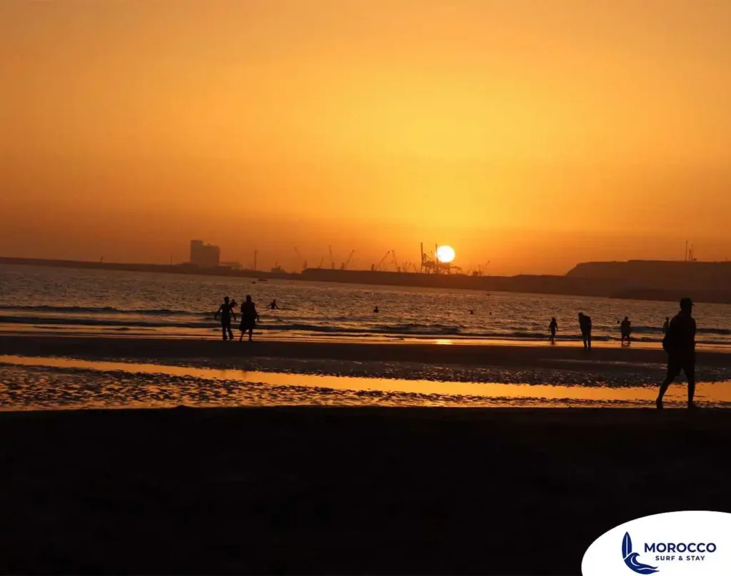 A beautiful sunset over Agadir beach, where the sky and wet sand are bathed in a brilliant orange and golden light. The silhouettes of several people are visible on the beach, including one person on horseback in the shallow water. In the distance, the sun is setting behind the hazy outline of the city's port, with its cranes visible against the horizon. The image captures a peaceful, end-of-day moment on the famous beach.