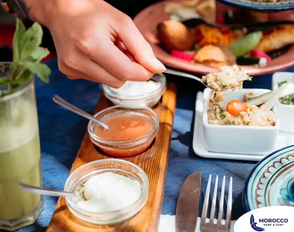 A close-up shot of a person's hand holding a spoon to scoop a creamy dip from a small white bowl. The table is set with a variety of Moroccan food, including small glass bowls of different sauces on a wooden tray and a mint drink on the side. The colorful dishes are arranged on a vibrant blue table.