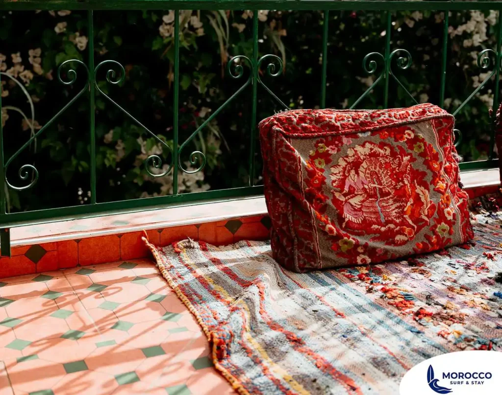 A close-up shot of a cozy corner on a tiled outdoor terrace in Morocco. A large, richly embroidered red floor cushion with a detailed pattern sits on a woven, colorful rug, creating a comfortable seating area next to a dark green metal railing. The background shows lush green plants with small white flowers, suggesting a vibrant and inviting space.