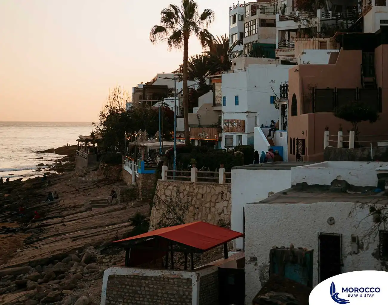 A serene, golden-hour view of the coastal village of Taghazout, Morocco. Terraced, white and earthy-toned buildings cascade down a rocky cliffside, with a prominent palm tree rising against the soft, glowing sky. The ocean is visible below, with waves gently breaking on the rocky shoreline where people are enjoying the sunset. The scene captures the quintessential charm of this famous surf village.