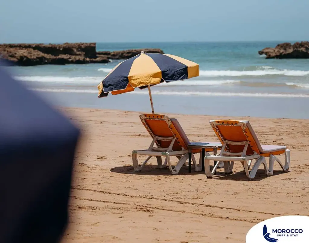 A peaceful view of two orange lounge chairs with a yellow and navy blue striped umbrella, set up on the golden sand of a beach in Tamraght, Morocco. The calm, blue ocean stretches out in the background, with large, rocky formations visible in the water to the left and right. The scene captures an inviting and relaxing moment on a sunny day.