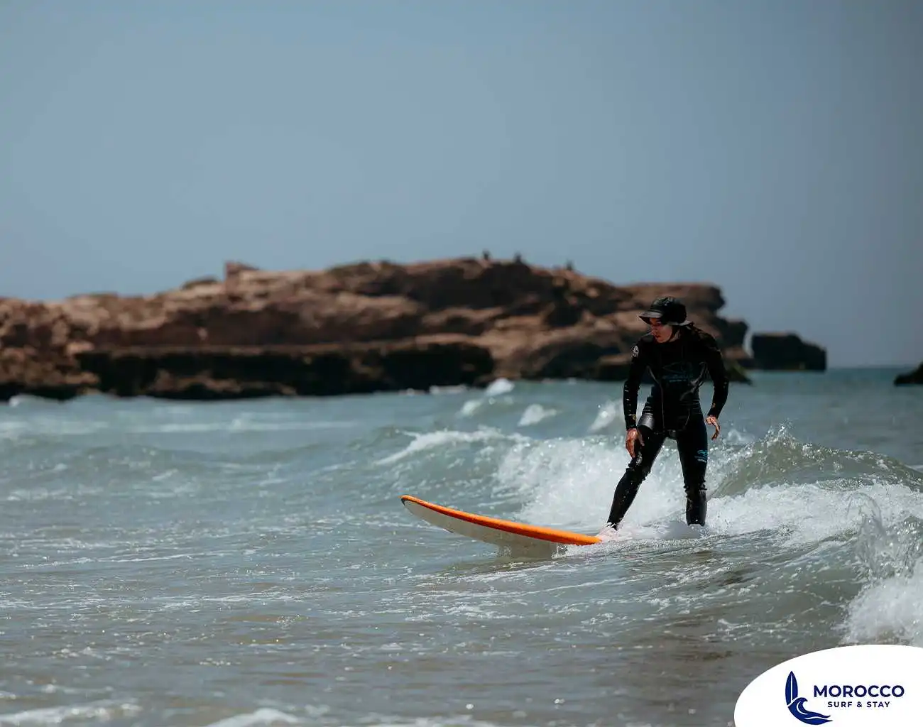 A surfer in a black wetsuit and a cap is skillfully riding a small wave on an orange surfboard in the waters off Tamraght, Morocco. The surfer is standing on the board, facing slightly to the left, with their arms outstretched for balance. In the background, there are rocky outcrops and the clear horizon under a bright sky, typical of this surfing destination.