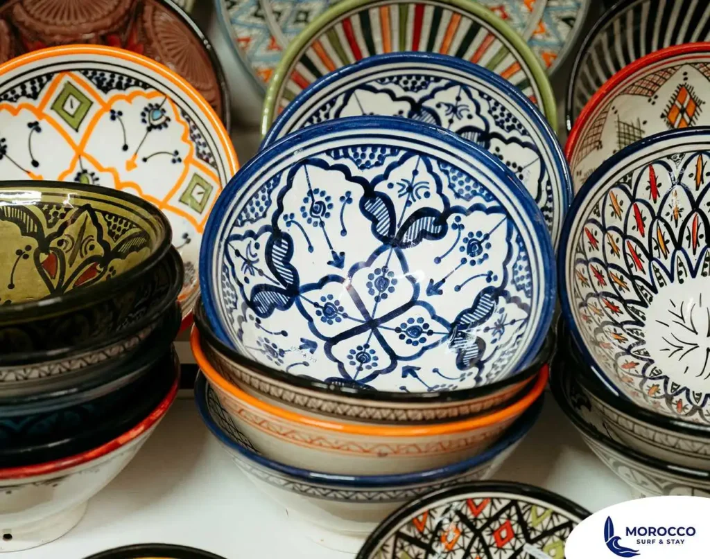 A close-up shot of a colorful display of handcrafted Moroccan pottery. In the foreground, a stack of bowls is topped with a white bowl featuring an intricate blue and black geometric pattern. Other stacks of plates and bowls with diverse and vibrant designs in shades of red, green, orange, and yellow are visible on either side, capturing the rich artistry and culture found in a Moroccan marketplace.