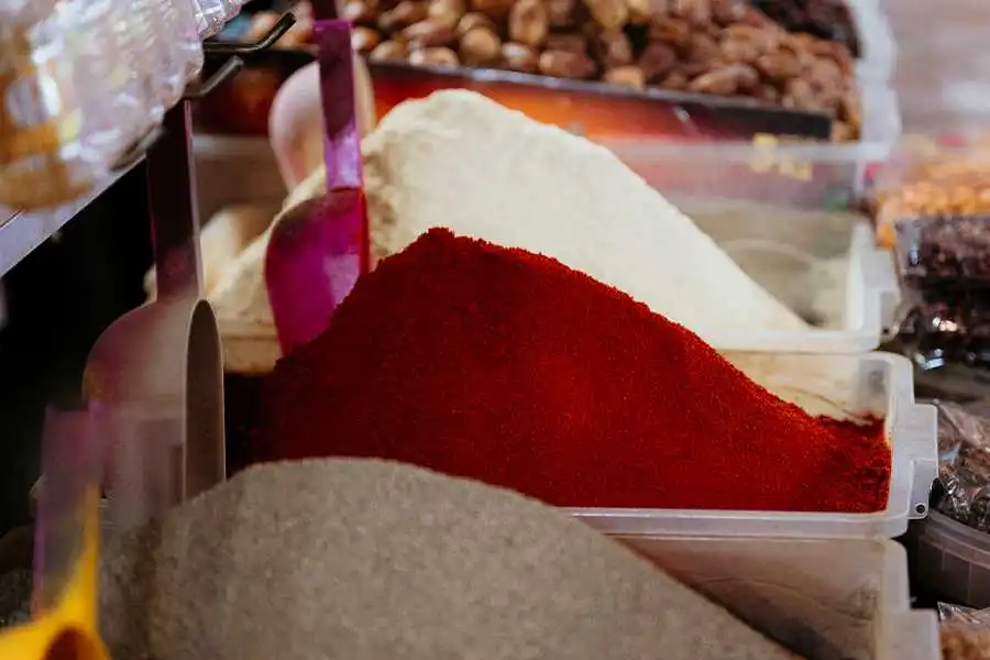 A close-up photograph shows various spices piled high in clear plastic containers at the Agadir Souk El Had market in Morocco. A large heap of vibrant red spice, likely paprika or chili powder, dominates the foreground. In the background, there are mounds of lighter-colored spices, possibly cumin or turmeric, along with nuts and dried fruits. A purple scoop is stuck into the red spice.
