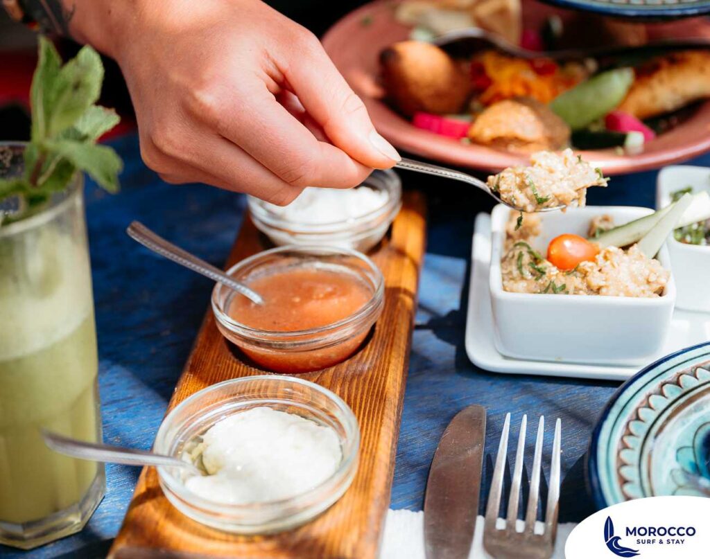 A close-up shot of a person's hand holding a spoon to scoop a creamy dip from a small white bowl. The table is set with a variety of Moroccan food, including small glass bowls of different sauces on a wooden tray and a mint drink on the side. The colorful dishes are arranged on a vibrant blue table.