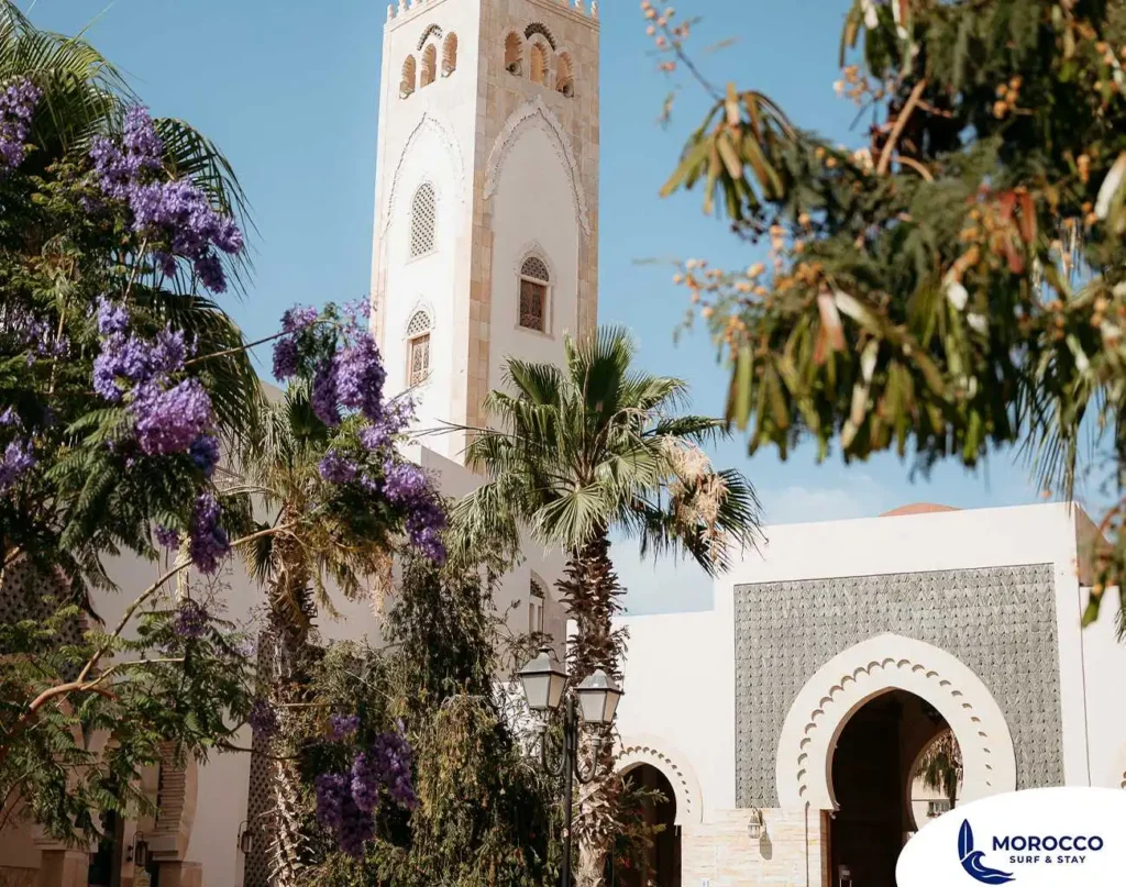 A low-angle exterior shot captures a beautiful, light-colored mosque with a tall, ornate minaret against a clear blue sky. The minaret features traditional Moroccan architectural details, including arched windows and decorative patterns. In the foreground and midground, vibrant purple jacaranda trees are in bloom, along with palm trees and other green foliage, framing the mosque and adding a pop of color to the scene. The lower part of the mosque also shows an arched entrance and decorative wall patterns.