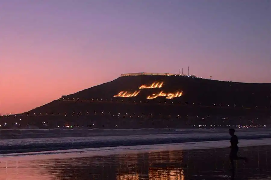 A wide, dusk shot of the coast of Agadir, with a person running along the wet sand in the foreground. The silhouette of a large, dark hill is visible in the distance, with the phrase 'الله، الوطن، الملك' (God, Homeland, King) written in lights on its side. The sky is a gradient of purple and orange, and lights from the city are visible below the hill.