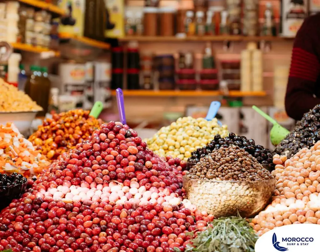A vibrant, close-up photograph of a variety of olives piled high at a stall in the Souk El Had market in Agadir, Morocco. The image shows large mounds of different kinds of olives, including shiny black, light green, and various shades of red and reddish-pink olives. Some of the piles have colorful scoops inserted into them. In the foreground, a large, clear bowl holds a mix of white and black olives. The background is a blur of other market stalls and goods.