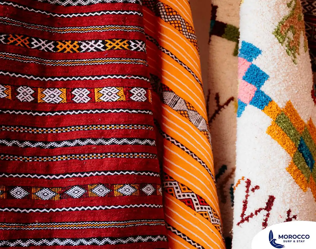 A close-up shot of several rolled-up Moroccan rugs or textiles, showcasing their rich colors and intricate patterns. The rug on the left is a vibrant red with black and white horizontal stripes and geometric designs. The middle rug is predominantly yellow and orange with a distinct tribal pattern. The rug on the right is white with a raised, colorful geometric pattern in shades of blue, pink, and orange.