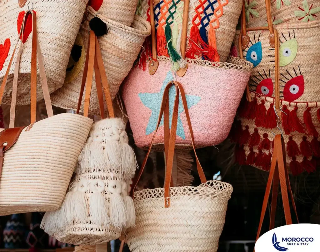 A close-up shot of an array of handcrafted straw bags and baskets hanging for sale at a market stall. The bags are made from woven straw and feature various embellishments, including colorful tassels, fringes, and embroidered designs such as a bright pink bag with a blue star and another bag with a face-like pattern. Several bags have brown leather handles.
