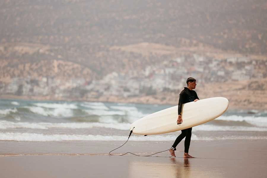 A male surfer in a black wetsuit walks along a wet, sandy beach in Taghazout, Morocco. He is carrying a long, white surfboard under his right arm. The background shows the ocean with gentle waves rolling in, and a hillside coastal town under a hazy sky, completing the classic surf town scene.