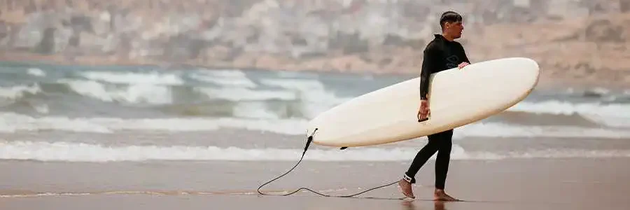 A full-length, side-profile shot of a man wearing a black wetsuit, walking along a wet, sandy beach while carrying a long, white surfboard under his right arm. A leash is attached to the board and his ankle. In the background, there are waves rolling onto the shore, and a hazy, mountainous coastline with buildings scattered across the hillsides. The overall scene has a soft, muted color palette.