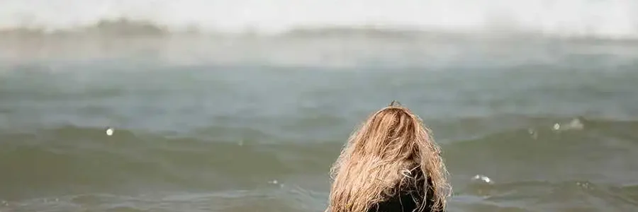 A young person with long, wavy blonde hair is sitting on a light blue surfboard in the shallow ocean water, facing away from the camera. They are wearing a black wetsuit. In the background, two other blurred surfers are visible further out in the water where a small wave is breaking.