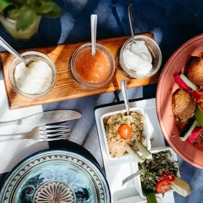 A close-up shot of a person's hand holding a spoon to scoop a creamy dip from a small white bowl. The table is set with a variety of Moroccan food, including small glass bowls of different sauces on a wooden tray and a mint drink on the side. The colorful dishes are arranged on a vibrant blue table.