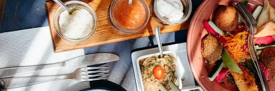 A close-up shot of a person's hand holding a spoon to scoop a creamy dip from a small white bowl. The table is set with a variety of Moroccan food, including small glass bowls of different sauces on a wooden tray and a mint drink on the side. The colorful dishes are arranged on a vibrant blue table.