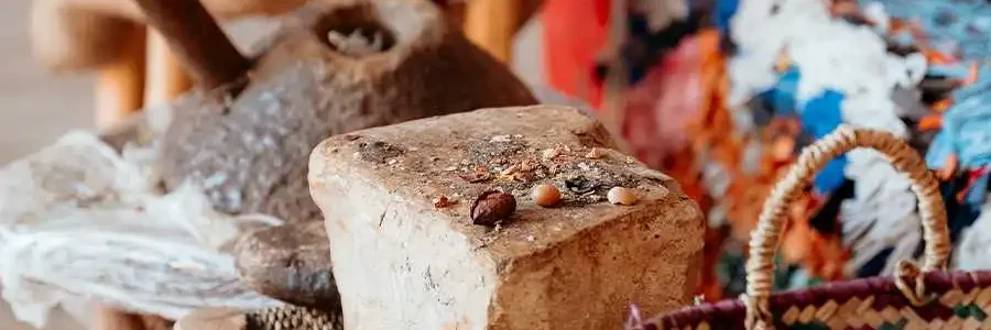 A close-up shot of various Moroccan artifacts in a market setting. The foreground shows a woven basket and a large, rough block of solid argan paste with several argan nuts on top. Behind it, a large, rustic pestle is visible, and to the side, a colorful woven rug or textile is partially shown. The lighting suggests an indoor or covered market space.