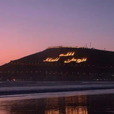 A wide, dusk shot of the coast of Agadir, with a person running along the wet sand in the foreground. The silhouette of a large, dark hill is visible in the distance, with the phrase 'الله، الوطن، الملك' (God, Homeland, King) written in lights on its side. The sky is a gradient of purple and orange, and lights from the city are visible below the hill.