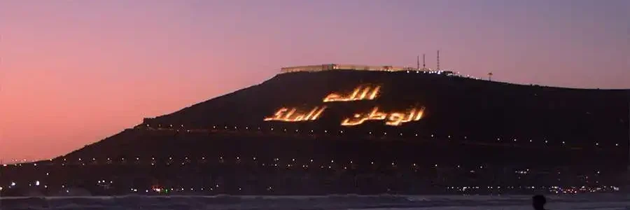 A wide, dusk shot of the coast of Agadir, with a person running along the wet sand in the foreground. The silhouette of a large, dark hill is visible in the distance, with the phrase 'الله، الوطن، الملك' (God, Homeland, King) written in lights on its side. The sky is a gradient of purple and orange, and lights from the city are visible below the hill.