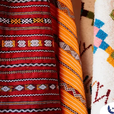 A close-up shot of several rolled-up Moroccan rugs or textiles, showcasing their rich colors and intricate patterns. The rug on the left is a vibrant red with black and white horizontal stripes and geometric designs. The middle rug is predominantly yellow and orange with a distinct tribal pattern. The rug on the right is white with a raised, colorful geometric pattern in shades of blue, pink, and orange.