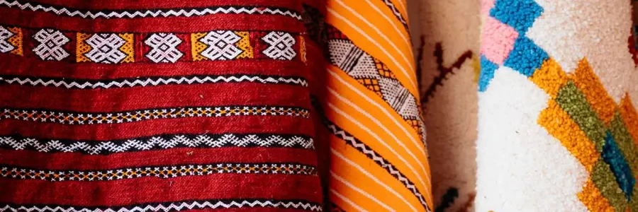A close-up shot of several rolled-up Moroccan rugs or textiles, showcasing their rich colors and intricate patterns. The rug on the left is a vibrant red with black and white horizontal stripes and geometric designs. The middle rug is predominantly yellow and orange with a distinct tribal pattern. The rug on the right is white with a raised, colorful geometric pattern in shades of blue, pink, and orange.