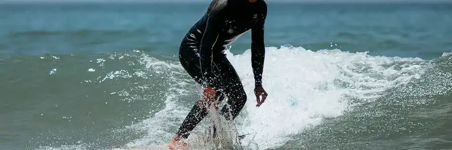 A close-up shot of a male surfer in a black wetsuit riding a small, foamy wave on a light-colored surfboard. He has dark, curly hair and a beard and is positioned in a low, balanced surfing stance. The water is a clear, vibrant blue, capturing a focused and dynamic moment of a surfer in action.
