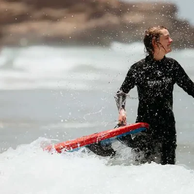 A young woman in a black wetsuit with the logo 'Darna Surf' is standing in the ocean, holding a red surfboard. She is smiling and looking to the side, with water splashing around her and the board. The background shows blurred waves and a rocky outcrop.
