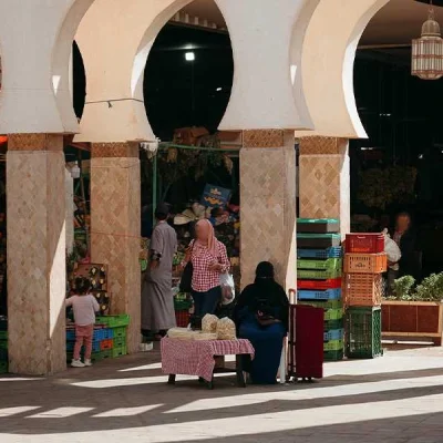 A bustling market scene under the traditional arched columns of Souk El Had in Agadir. The photo shows several people, including locals and vendors, among stalls filled with colorful fruits and vegetables in plastic crates. The bright sunlight casts shadows across the tiled ground, highlighting the lively atmosphere of the market.