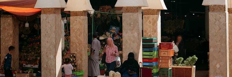 A bustling market scene under the traditional arched columns of Souk El Had in Agadir. The photo shows several people, including locals and vendors, among stalls filled with colorful fruits and vegetables in plastic crates. The bright sunlight casts shadows across the tiled ground, highlighting the lively atmosphere of the market.