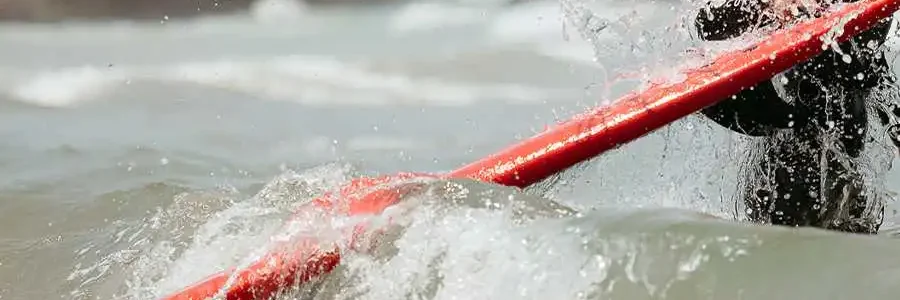 A close-up, dynamic shot of a surfer in a black wetsuit holding a bright red surfboard. The surfer is standing in the ocean as a wave crashes and splashes around the board and their body, creating a lot of white spray. A rocky coastline is blurred in the background.