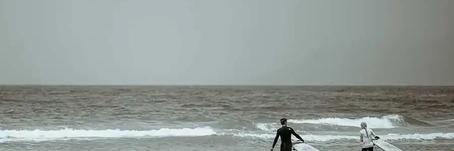 A sepia-toned photograph showing two surfers, one in a dark wetsuit and one in a light top, walking into the ocean with their surfboards. They are partially submerged in the shallow waves, with more waves further out in the sea under a hazy sky.