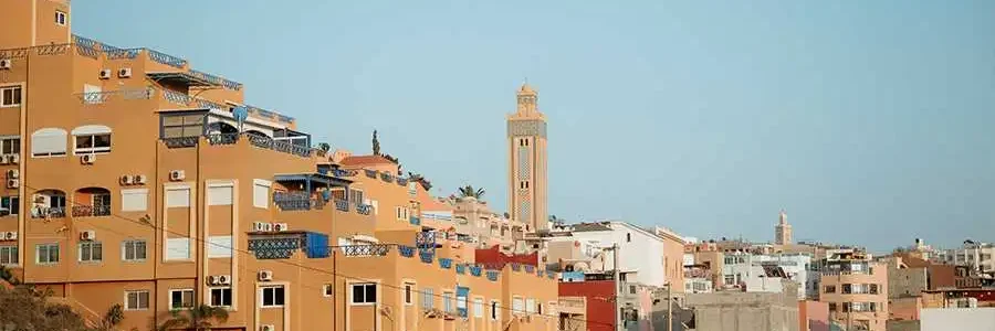 A wide shot of a Moroccan hillside town. The foreground is a dry, rocky slope with sparse vegetation. The mid-ground is dominated by orange and light-colored buildings with balconies and small windows. In the background, a tall, yellow-beige minaret with geometric patterns and a smaller one in the distance rise above the other rooftops against a clear blue sky.
