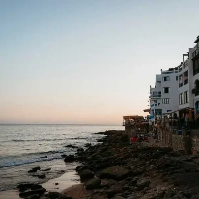 A wide shot of the Taghazout coastline at sunset. On the right, a row of white multi-story buildings and restaurants with arched balconies sits on a rocky shoreline. The foreground shows a mix of sand and dark rocks with small waves lapping at the shore. The horizon features a smooth transition from the pale blue sky to a light orange glow as the sun sets over the calm ocean.