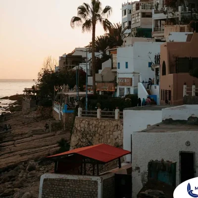 A serene, golden-hour view of the coastal village of Taghazout, Morocco. Terraced, white and earthy-toned buildings cascade down a rocky cliffside, with a prominent palm tree rising against the soft, glowing sky. The ocean is visible below, with waves gently breaking on the rocky shoreline where people are enjoying the sunset. The scene captures the quintessential charm of this famous surf village.