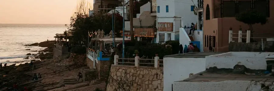 A serene, golden-hour view of the coastal village of Taghazout, Morocco. Terraced, white and earthy-toned buildings cascade down a rocky cliffside, with a prominent palm tree rising against the soft, glowing sky. The ocean is visible below, with waves gently breaking on the rocky shoreline where people are enjoying the sunset. The scene captures the quintessential charm of this famous surf village.