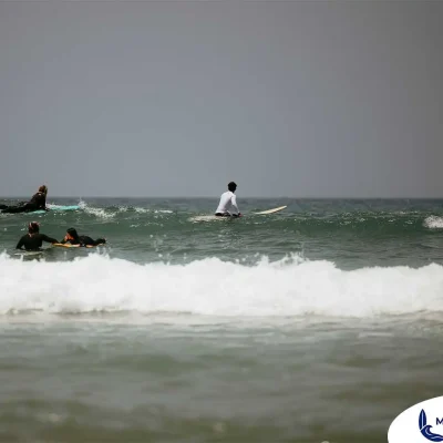 A group of surfers is seen during a lesson in the waters of Taghazout, Morocco. In the foreground, a white-capped wave rolls towards the shore, while four people on surfboards, including one in a white shirt and others in black wetsuits, are spread out beyond it, either paddling or waiting for a wave. The image captures the active scene of learning to surf at this famous beach.