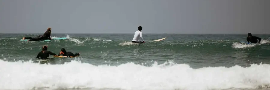 A group of surfers is seen during a lesson in the waters of Taghazout, Morocco. In the foreground, a white-capped wave rolls towards the shore, while four people on surfboards, including one in a white shirt and others in black wetsuits, are spread out beyond it, either paddling or waiting for a wave. The image captures the active scene of learning to surf at this famous beach.