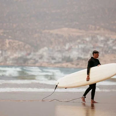 A male surfer in a black wetsuit walks along a wet, sandy beach in Taghazout, Morocco. He is carrying a long, white surfboard under his right arm. The background shows the ocean with gentle waves rolling in, and a hillside coastal town under a hazy sky, completing the classic surf town scene.