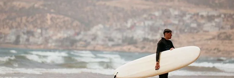 A male surfer in a black wetsuit walks along a wet, sandy beach in Taghazout, Morocco. He is carrying a long, white surfboard under his right arm. The background shows the ocean with gentle waves rolling in, and a hillside coastal town under a hazy sky, completing the classic surf town scene.