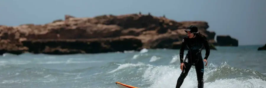 A surfer in a black wetsuit and a cap is skillfully riding a small wave on an orange surfboard in the waters off Tamraght, Morocco. The surfer is standing on the board, facing slightly to the left, with their arms outstretched for balance. In the background, there are rocky outcrops and the clear horizon under a bright sky, typical of this surfing destination.